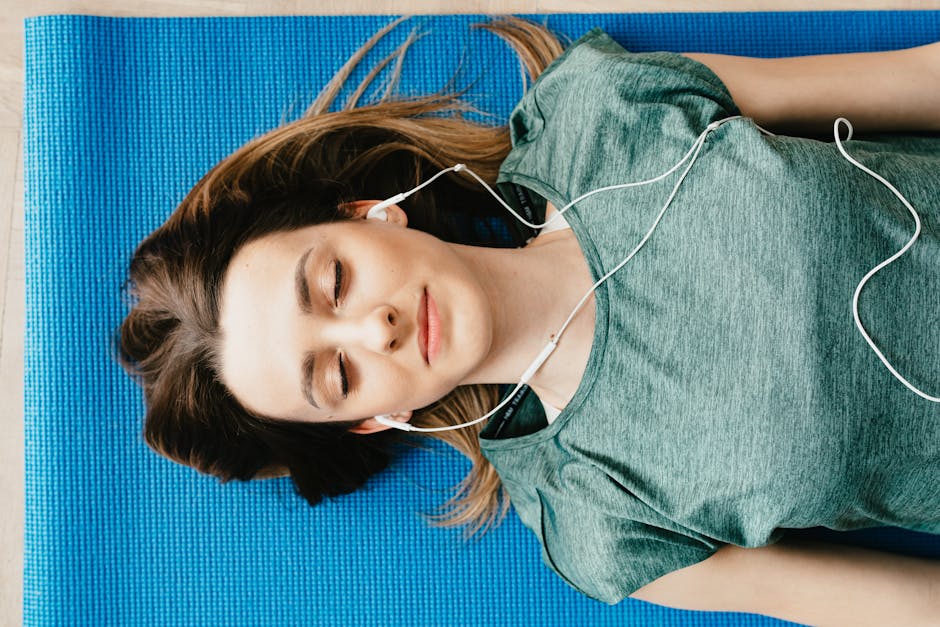 From above of calm young female in casual wear listening to music in earphones while taking break after workout on blue mat in flat