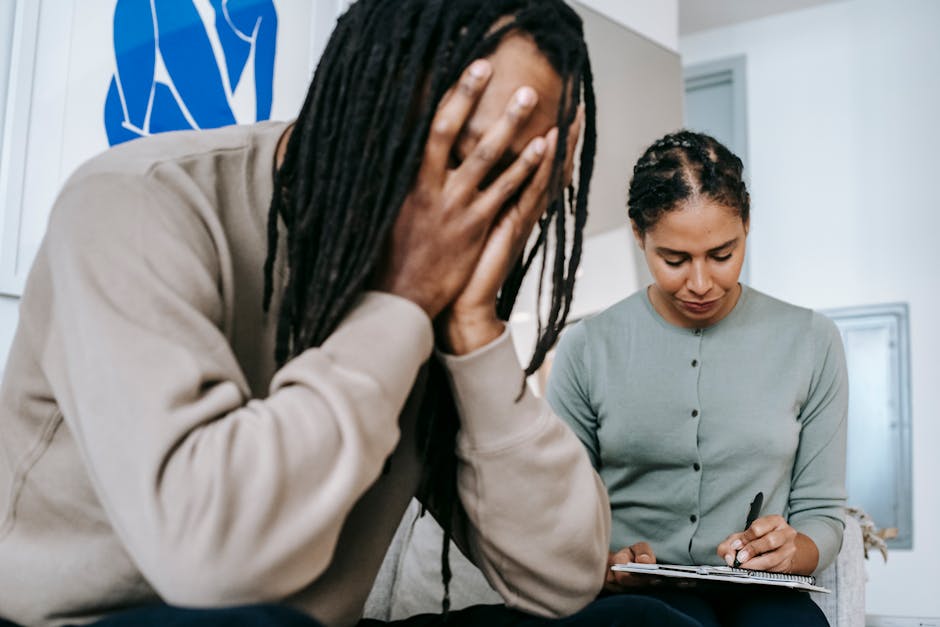 African American man with dreadlocks covering face with hands and sitting near Hispanic woman writing notes in during therapy session