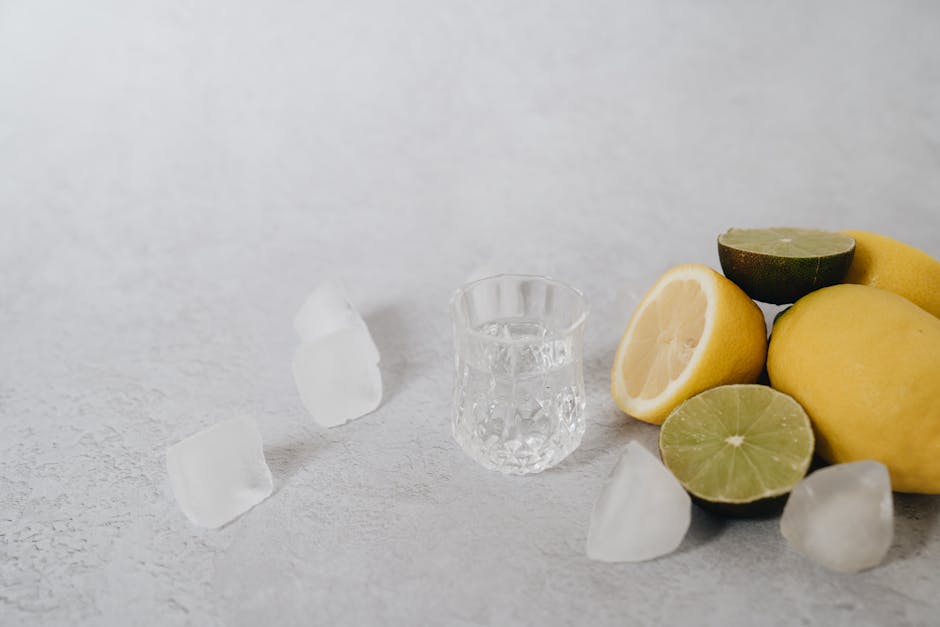Fresh lemons and limes on a textured surface with ice cubes and a glass of water.